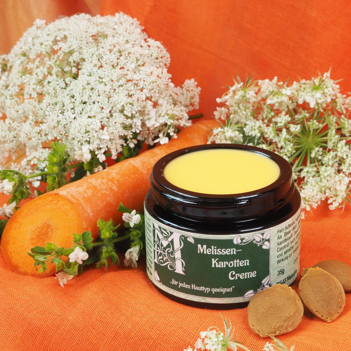 Geöffneter Melissen-Karotten Creme Tiegel 35ml. Daneben liegt eine Karotte.