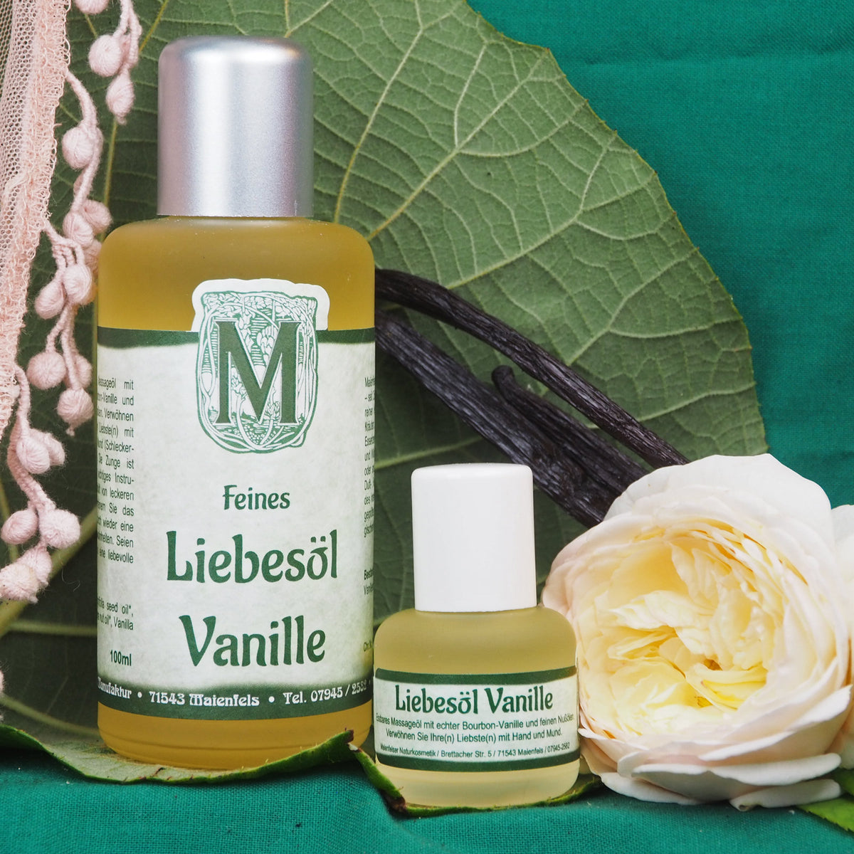 Liebesöl Vanille abgefüllt in Glasflasche jeweils in der Größe 100ml und 15ml. Im Hintergrund eine helle Rose mit großem grünem Blatt