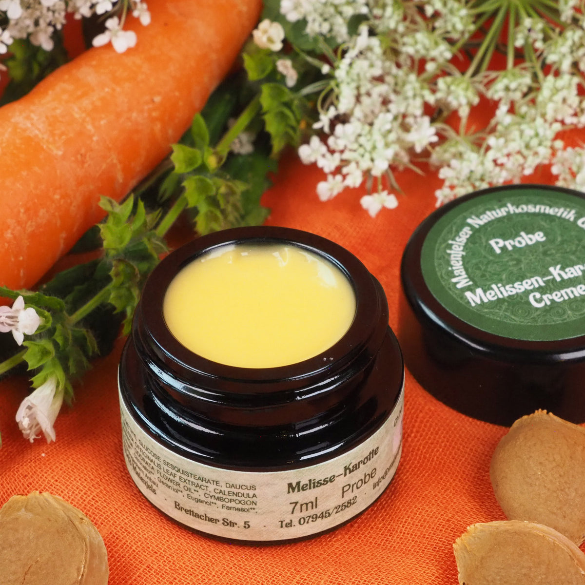 Geöffneter 7ml Melissen-Karotten Creme Tiegel, Daneben liegt der Deckel der Creme und eine Karotte.